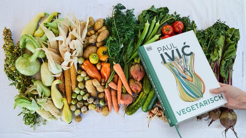 Auf einem Tischtuch ist eine Vielfalt unterschiedlicher Gemüse farblich sortiert aufgelegt. Darüber hält eine Hand das Kochbuch "Vegetarisch" von Paul Ivić in die Kamera.