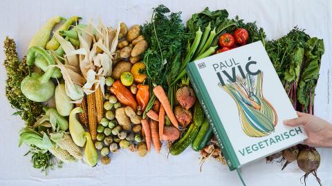 Auf einem Tischtuch ist eine Vielfalt unterschiedlicher Gemüse farblich sortiert aufgelegt. Darüber hält eine Hand das Kochbuch "Vegetarisch" von Paul Ivić in die Kamera.